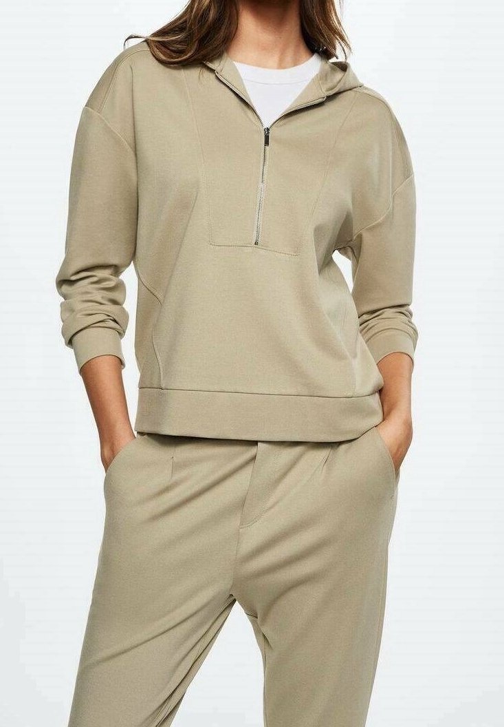 Femme portant un sweat-shirt beige à demi-fermeture éclair et un pantalon assorti, les mains dans les poches, sur un fond uni.