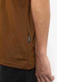 Hnědá bavlněná košile s krátkým rukávem, která zobrazuje boční šev s tkanou etiketou. Texturovaná tkanina, uvolněný střih a mírně zakřivený lem.