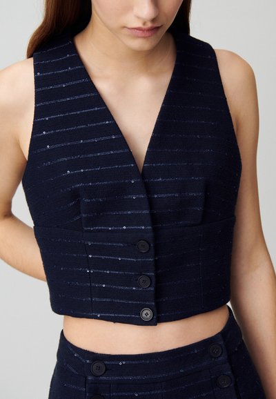 Veste noire raccourcie avec des rayures verticales, sequins accentuant le tissu, décolleté en V profond et cinq boutons noirs pour la fermeture.