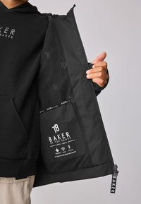 Osoba drží otvorenú čiernu bundu, ktorá ukazuje vnútorné vrecko s logom "Baker by Ted Baker" a ikonami nepremokavosti, odvodnenia a odolnosti voči búrkam.