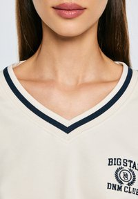 Beige T-Shirt mit V-Ausschnitt und navyfarbigem Rippkragen, auf der linken Seite mit einem Logo und Lorbeerdesign, aus glattem Stoff gefertigt.