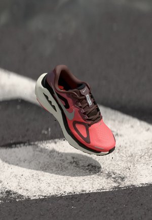 Αθλητικό παπούτσι Nike με ροζ και καφέ επάνω μέρος από δίχτυ, μαύρη υποστήριξη στη φτέρνα και λευκή μεσαία σόλα, τοποθετημένο πάνω σε βαμμένη επιφάνεια.