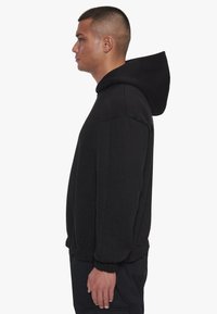 Schwarzer Kapuzenpullover mit lockerem Schnitt, gerippten Bündchen und Saum, aus einem strukturierten Stoff und mitSeitentaschen, seitlich präsentiert.