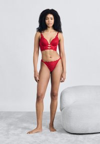 Κόκκινο δαντελένιο σουτιέν και στρινγκ που ταιριάζει με βαθύ ντεκολτέ, κυματιστά άκρα και διακοσμητικές εγκοπές στο σουτιέν. Το μοντέλο στέκεται ξυπόλυτο.