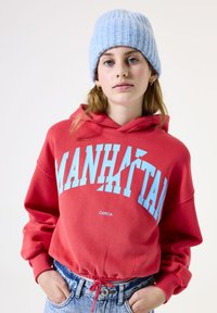 Mujer joven vistiendo una sudadera roja "Manhattan" y un gorro de lana azul claro, de pie con las manos en los bolsillos de unos jeans azules contra un fondo blanco.