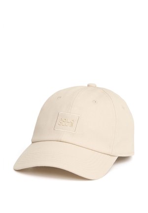 Gorra de béisbol beige con visera curva y un pequeño parche cuadrado en la parte frontal que muestra el texto en relieve "POLA CAVE".