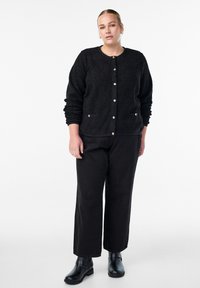 Sieviete, tērpusies melnā teksturētā jaku ar pogām, melnās plašās biksēs un melnās potīšu zābakos, stāv uz baltas fona.