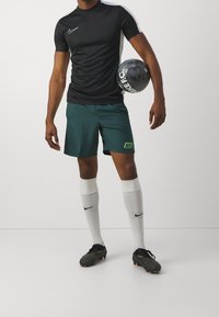 Hombre con camiseta deportiva negra de Nike, pantalones cortos verdes de Nike, calcetines blancos de Nike y zapatos deportivos negros, sosteniendo un balón de fútbol negro de Nike bajo el brazo.