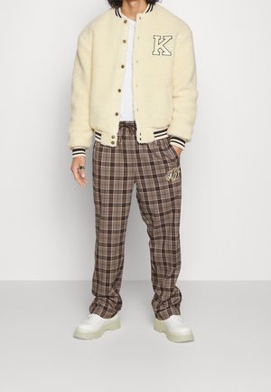 Person trägt eine cremefarbene Collegejacke mit dem Buchstaben K, ein weißes Hemd, braune karierten Jogginghosen mit goldenen Initialen und weiße Plateauschuhe.