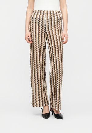 Donna che indossa pantaloni a gamba larga a vita alta con motivo a zigzag beige, marrone e nero, abbinati a décolleté nere con punta aguzza.