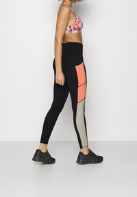 Leggings neri con blocchi di colore rosa e beige, design aderente e texture liscia. Abbinati a un reggiseno sportivo multicolore e scarpe da ginnastica nere.