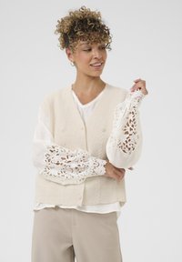 Kreminė raukinėta liemenė su perlų mygtukais ant baltos palaidinės, kurioje yra nėrinių detalės ant rankovių. Minkšta tekstūra, sluoksniuotas įvaizdis, neutrali spalvų paletė.