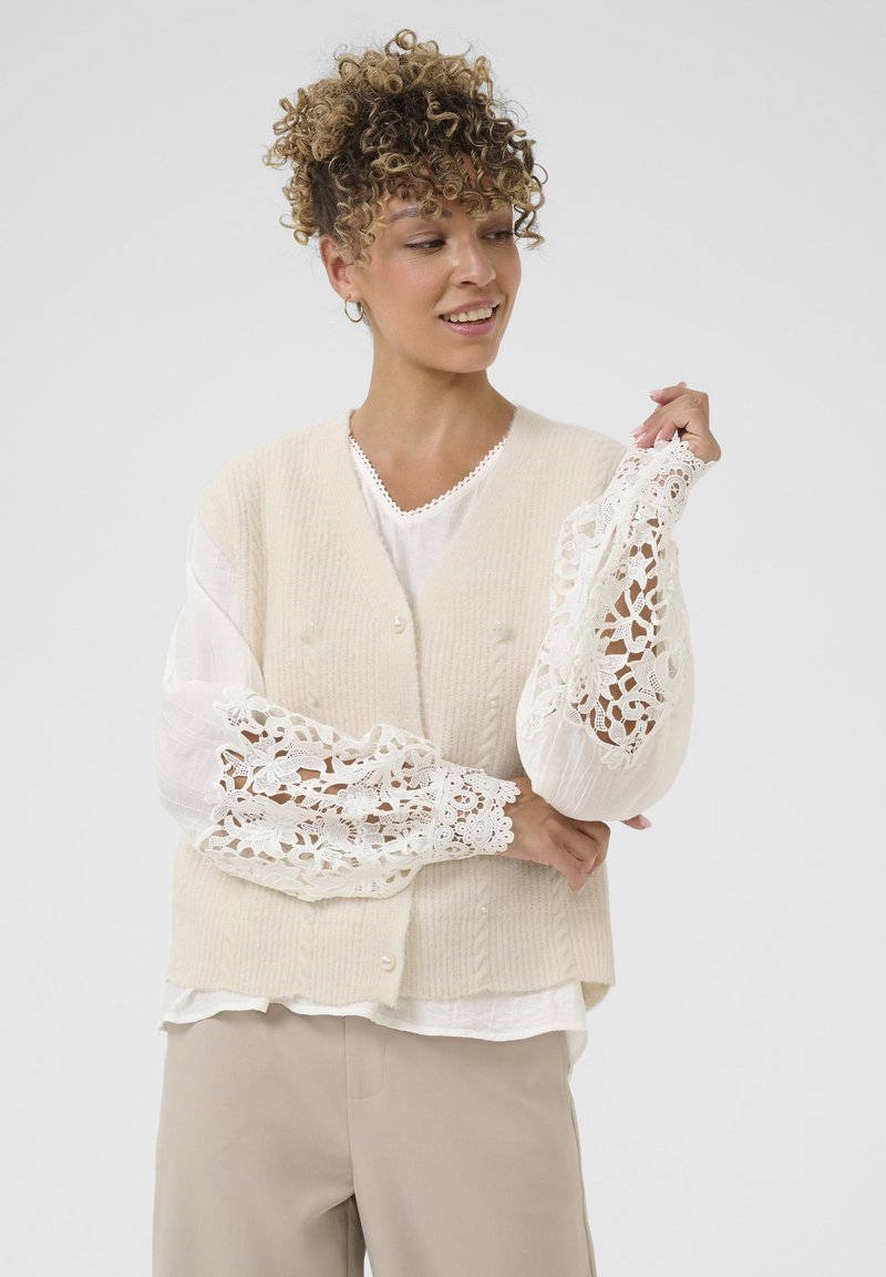 Kreminė raukinėta liemenė su perlų mygtukais ant baltos palaidinės, kurioje yra nėrinių detalės ant rankovių. Minkšta tekstūra, sluoksniuotas įvaizdis, neutrali spalvų paletė.