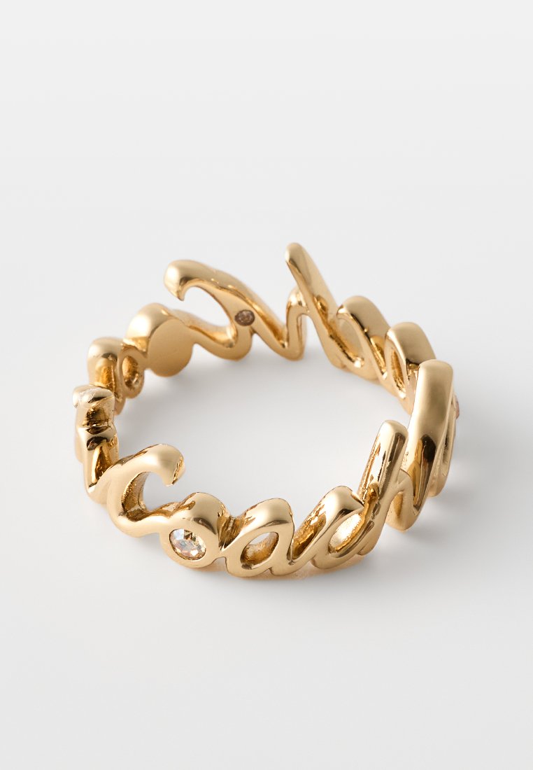 Goldfarbener Ring in Schreibschrift mit den Wörtern "Engel" und "Skarabäus" sowie kleinen eingearbeiteten klaren Edelsteinen auf weißem Hintergrund.
