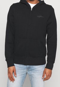 Musta vetoketjullinen huppari, valmistettu pehmeästä puuvillamateriaalista, jossa edessä tasku ja huppu. Rintakehällä on hillitty harmaa logotunnus.