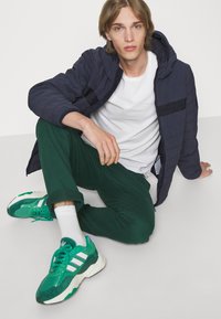 Giacca a cappuccio trapuntata navy, t-shirt bianca, pantaloni verdi, sneakers verdi con strisce bianche e logo, seduto su uno sfondo semplice.