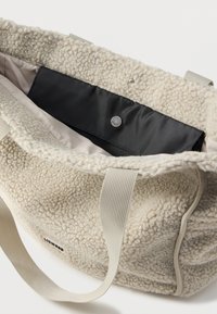 Beige υφασμάτινη τσάντα tote με μαλακή υφή, διπλές υφασμάτινες λαβές, εσωτερική μαύρη επένδυση, κλείσιμο με κουμπί και ταμπελάκι με λογότυπο στο πλάι.