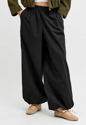 Personne portant un pantalon noir ample avec des poignets élastiques, des ballerines beige avec des rubans noués, un haut blanc court et une veste vert olive.