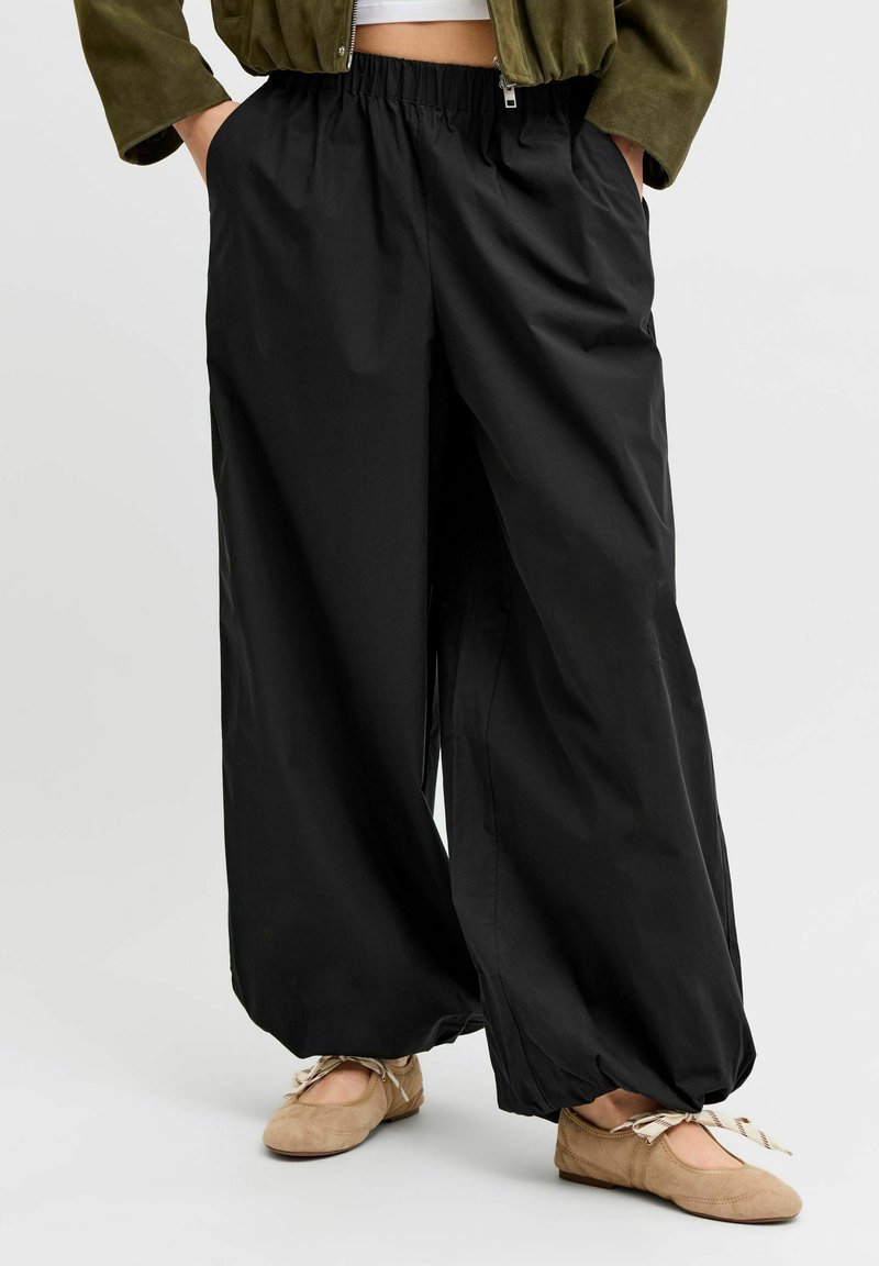 Personne portant un pantalon noir ample avec des poignets élastiques, des ballerines beige avec des rubans noués, un haut blanc court et une veste vert olive.