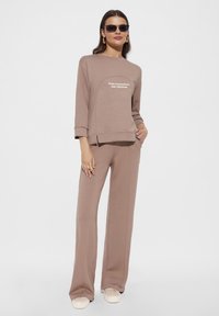 Brązowy, długi rękaw, bluza z tekstem "URODZONY Z PASSJĄ I WOLNOŚCIĄ" w zestawie z dopasowanymi spodniami typu wide-leg. Model w okularach przeciwsłonecznych i sneakersach.