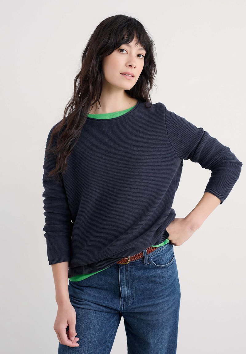 Pull en maille côtelée bleu marine foncé avec une doublure verte. Manches longues, coupe décontractée, porté avec un jean bleu clair et une ceinture tressée marron.