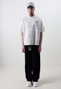 White short-sleeve t-shirt with layered pleats, black loose pants, and white sneakers. Gray cap worn. Minimalist design with logo detail.