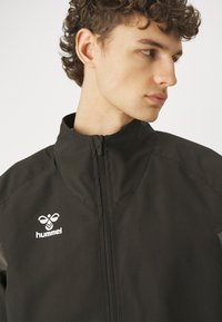 Svart vindjacka med hög krage, full dragkedja och vit logo på vänster bröst. Slät tyg med en något glänsande yta.