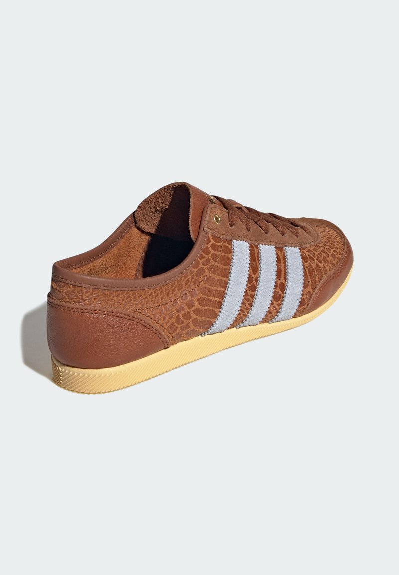 Brune sneakers med en tekstureret overlæder, der ligner reptilskind, hvide striber og en gul gummisål. Har brune snørebånd og gyldne snørehuller.