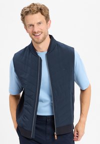 Marine gewatteerde vest met hoge kraag en ritssluiting, gecombineerd met een lichtblauwe blouse met korte mouwen, voorzien van een geribbelde onderrand en zijkantaccenten.