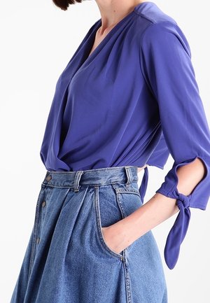 Vrouw draagt een paarse wikkelblouse met gestrikte mouwen en een hoog uitgesneden spijkerrok met knopen en zakken, hand in de zak gestoken.