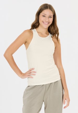 Ung kvinde smilende iført en ærmeløs cremefarvet top og løse beige bukser, poserer med den ene hånd på hoften foran en ensfarvet baggrund.