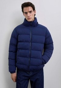 Hombre con una chaqueta acolchada de color azul marino, de cuello alto, con cremallera frontal y puños elásticos, de pie contra un fondo claro y liso.