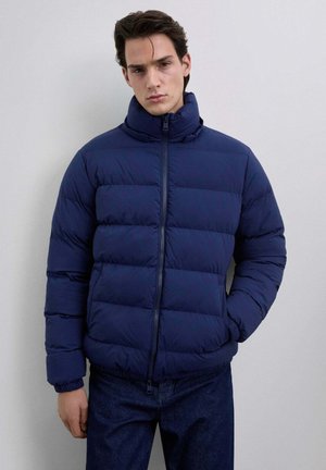 Hombre con una chaqueta acolchada de color azul marino, de cuello alto, con cremallera frontal y puños elásticos, de pie contra un fondo claro y liso.