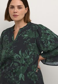 Blusa scura con motivi floreali verdi, scollatura a v e maniche a palloncino. Realizzata in un tessuto morbido con una finitura strutturata.