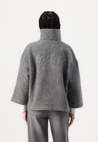 Jersey de lana gris oversize con cuello alto y logotipo bordado en la espalda. Presenta mangas anchas y una textura suave. Combinado con jeans grises.