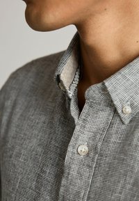 Grå skjorte med knaplukning og et struktureret, rutenet mønster. Indersiden af kraven har et kontrasterende lyst stof med et subtilt design. To synlige knapper.