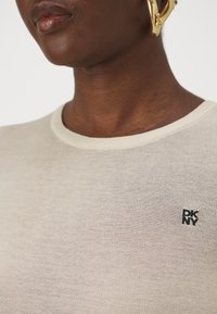 Beige strikket genser med rund hals og svart DKNY-logo på brystet. Myk tekstur, subtilt mønster og tettsittende design.