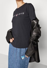 Person, der bærer en sort GANT 1949 t-shirt, en blå denim-nederdel med en trekantet lap, og holder en sort læderjakke hængende over den ene arm.