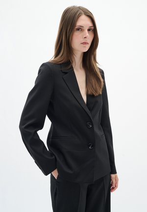 Mladá žena s dlhými hnedými vlasmi, ktorá nosí čierne na mieru šité sako a zodpovedajúce nohavice, stojí s jednou rukou v vrecku na bielom pozadí.