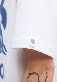 Hvid bomulds t-shirt med et blåt grafisk design. Ærmet har et rektangulært mærke med "HUGO" og et logo i grå.