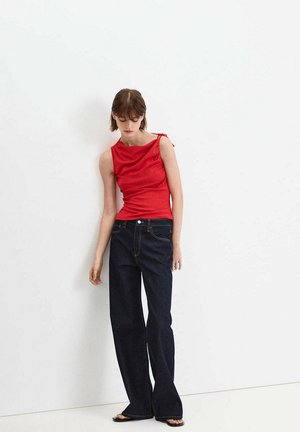 Giovane donna con top rosso senza maniche e jeans scuri a gamba larga, in piedi a piedi nudi contro un muro bianco, guarda in basso.