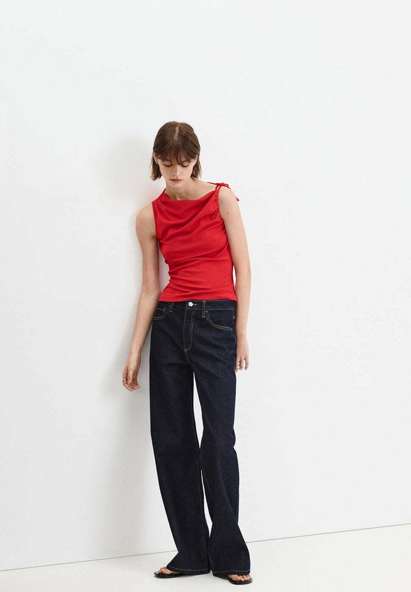 Jeune femme en haut rouge sans manches et jean large foncé, debout pieds nus contre un mur blanc, regardant vers le bas.
