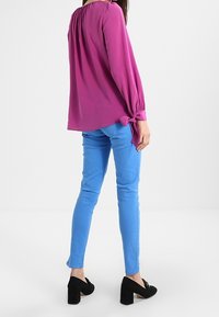 Blusa fucsia de manga larga con escote fruncido y detalle de lazo, combinada con jeans ajustados de color azul claro y zapatos de tacón negros.