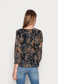 Sort bluse med blomsterprint i blå og orange, med lange ærmer, en nøglehulslukning bagpå og en afslappet pasform. Matchet med blå jeans.