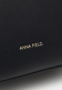 Černý texturovaný materiál s embosovaným logem "ANNA FIELD" ve zlaté barvě. Elegantní design s minimálním hardwarem a čistými liniemi, který zdůrazňuje moderní estetiku.