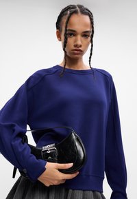 Sötétkék pulóver hosszú ujjakra, bordás nyakkal és mandzsettákkal. Fekete, textúrált kézitáska csattos részletekkel, amelyet egy szürke csíkozott szoknya mellett tartanak.