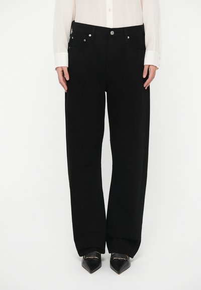 Pantalones negros de pierna ancha hechos de mezclilla, con cierre de botones, bolsillos delanteros y textura suave. Combinados con zapatos negros puntiagudos.
