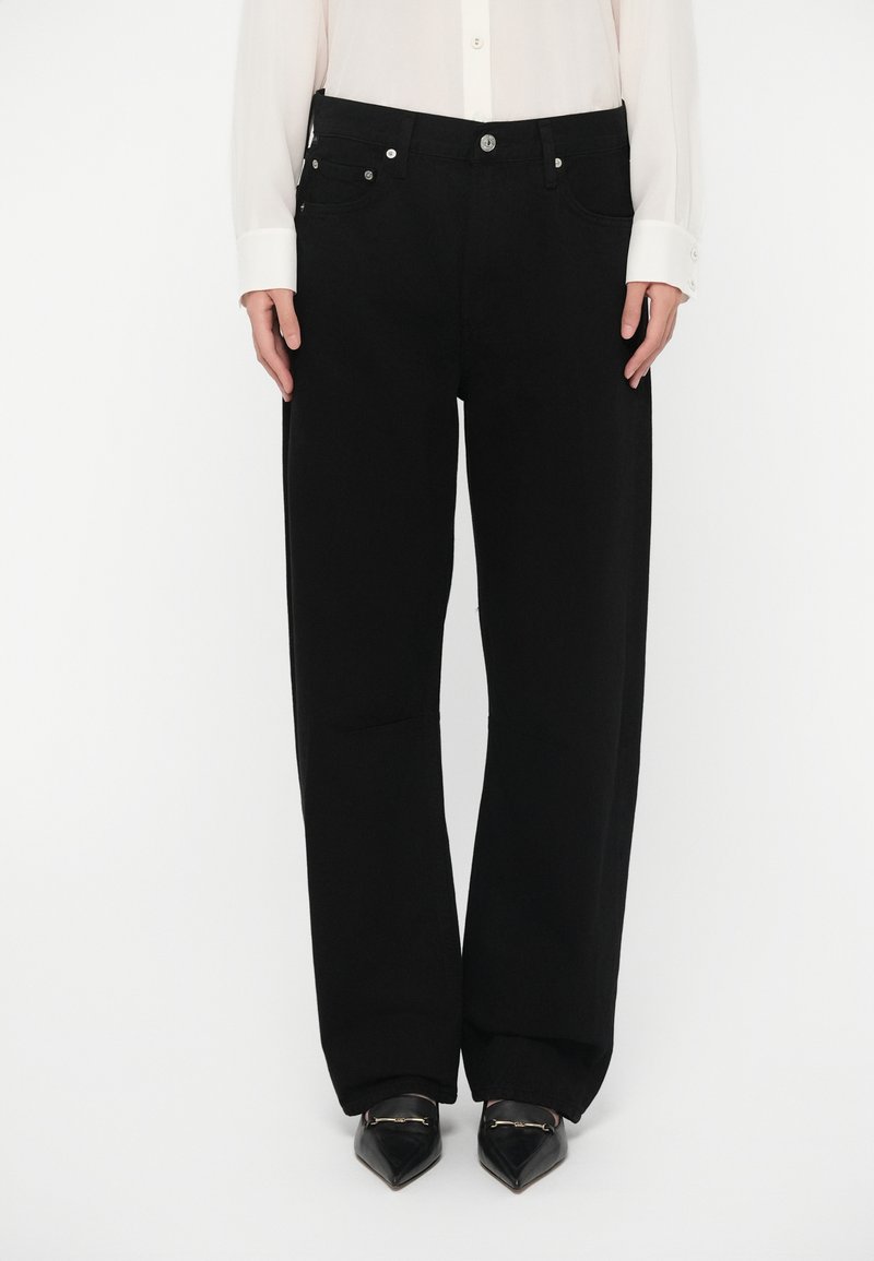 Melnas platām kājām džinsa bikses ar pogas aizdari, priekšējām kabatām un gludu faktūru. Valkātas ar melniem, asinātiem apaviem.