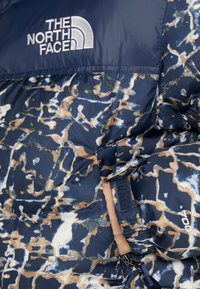 Veste North Face à motif camouflage bleu et marron avec logo visible, poignet avec sangle velcro, partie de fermeture éclair et main dans la manche.