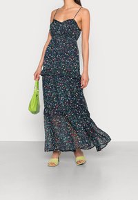 Floral maxi dress in navy with spaghetti straps, tiered design, and a lightweight texture. Paired with a green handbag and matching shoes.